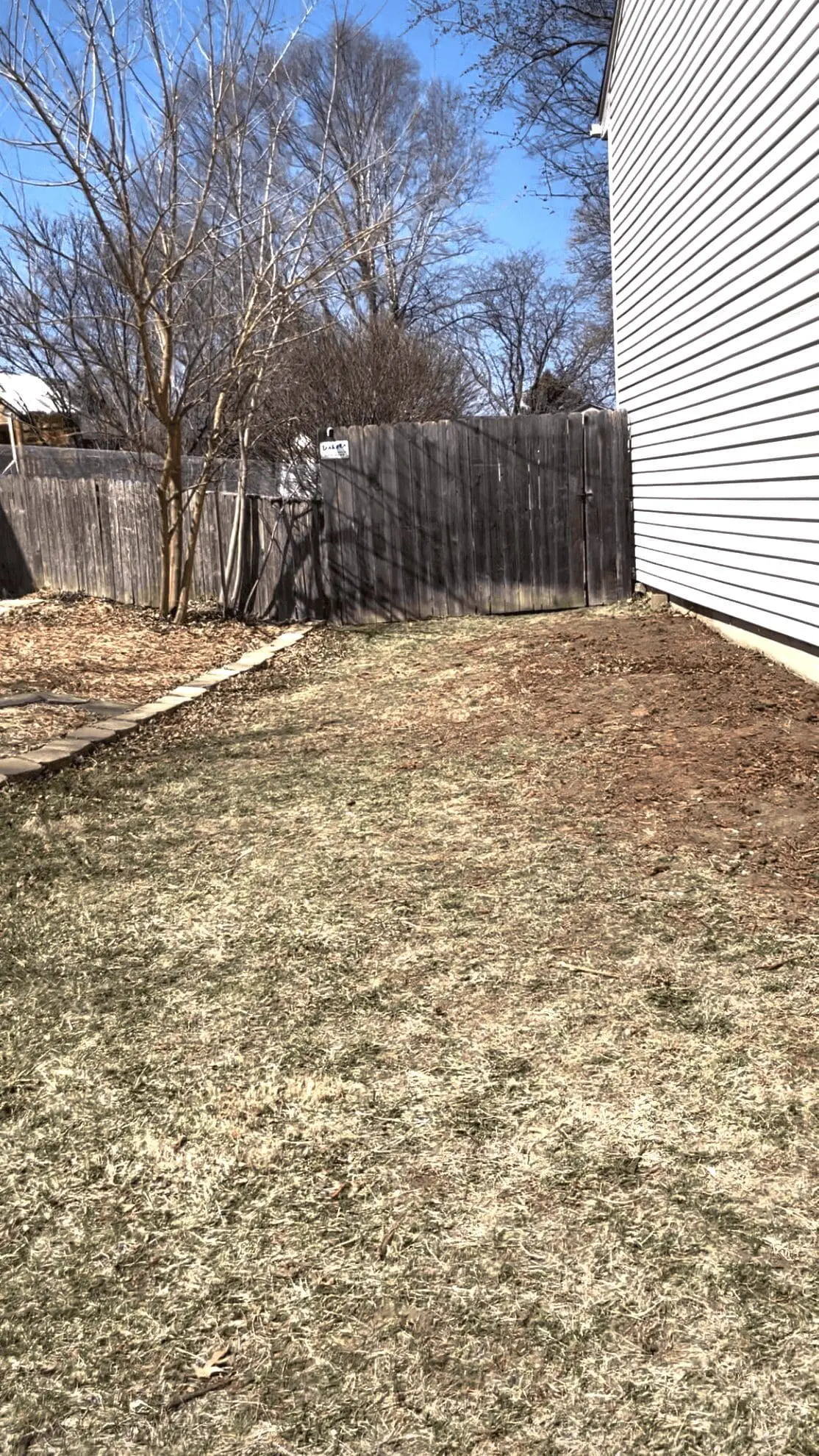 Side yard space with patchy grass and freshly turned soil beside a white house with vertical vinyl siding. A wooden privacy fence and leafless trees are in the background, hinting at early spring.